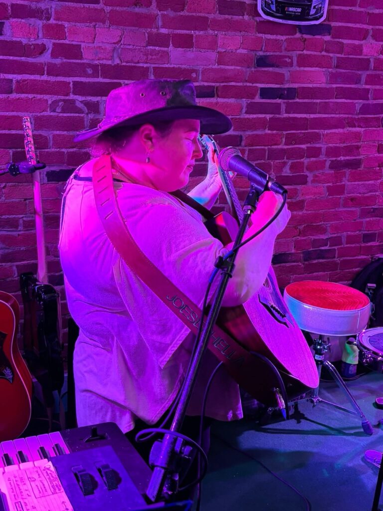 Bobbie JoEle playing guitar in a bar, lit from behind by red stage lights